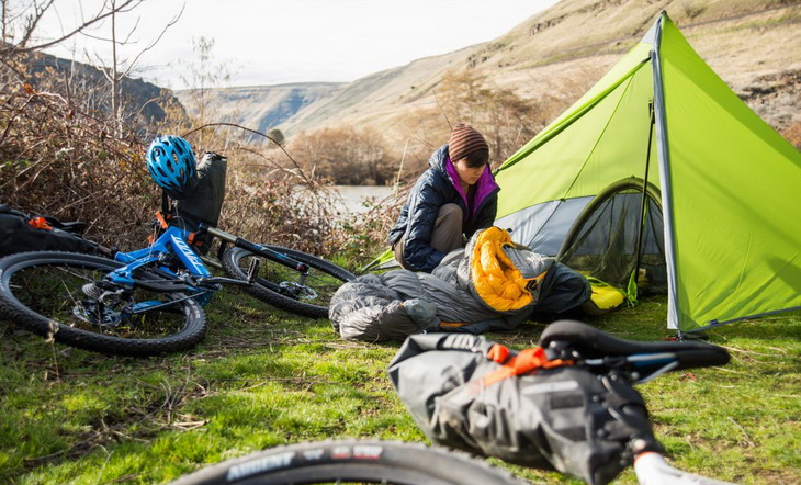 Woman looking down to Nemo sleeping bag sitting near two bikes and a tent