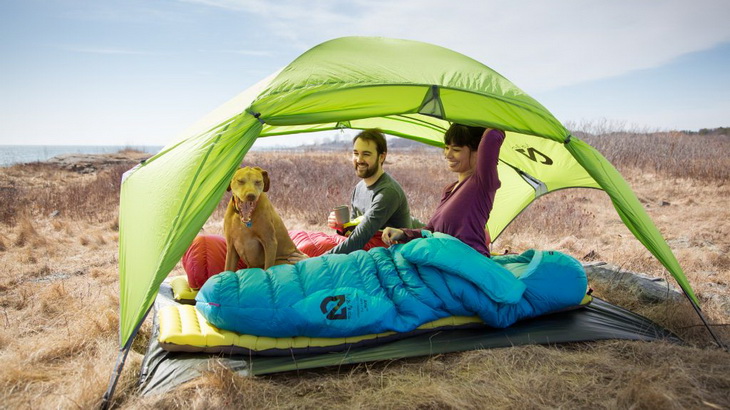 Two adults in Nemo sleeping bags next to a dog sitting in a tent