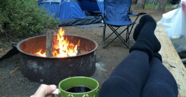A woman relaxing with a cup of coffee in the camp near a fire and a tent