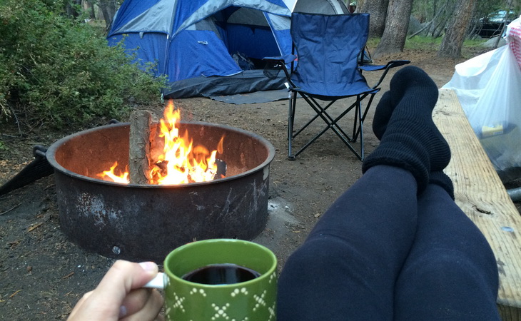 A woman relaxing with a cup of coffee in the camp near a fire and a tent