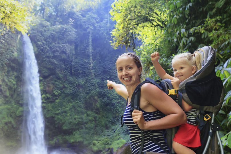 Mother hold baby girl in backpack on waterfall background
