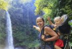 Mother hold baby girl in backpack on waterfall background