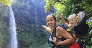 Mother hold baby girl in backpack on waterfall background