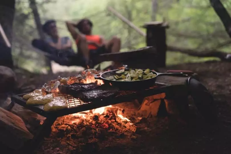 Black Non Stick Pan on Black Metal Charcoal Griller With Steak on Outdoor Forest With Two Persons Seating on Hammock