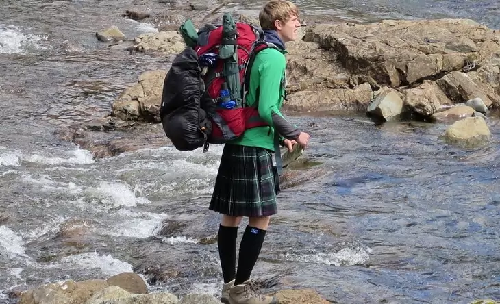 A man with a backpack and wearing a kilt trying to pass the river