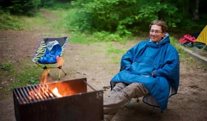Man wearing a blue poncho is sitting on a chair near a camp fire