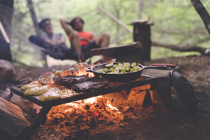 Black Non Stick Pan on Black Metal Charcoal Griller With Steak on Outdoor Forest With Two Persons Seating on Hammock