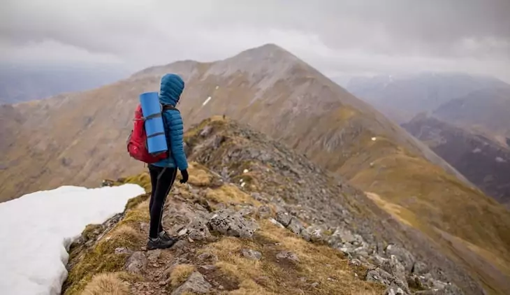 Person in Blue Hoodie Jacket Wearing Red Hiking Backpack Standing at the Top of Mountain Under White Sky during Daytime