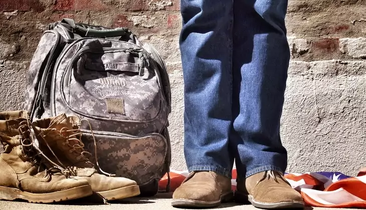 Person in Blue Jeans and Brown Suede Shoes Standing Near Camouflage Backpack Brown Hiking Boots and American Flag on Floor