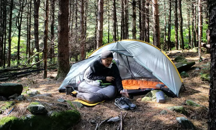 A man in Nemo Siren Down Sleeping Bag laying down near a tent in the forest