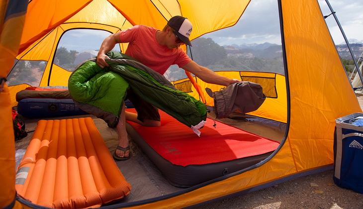 Man in a tent holding a sleeping bag and a pair of pants
