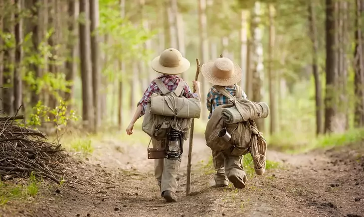 Two boys go hiking with backpacks on a forest road bright sunny day