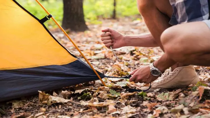 Man in plaid shirt and sneakers crouches on leaves to push tent peg into ground