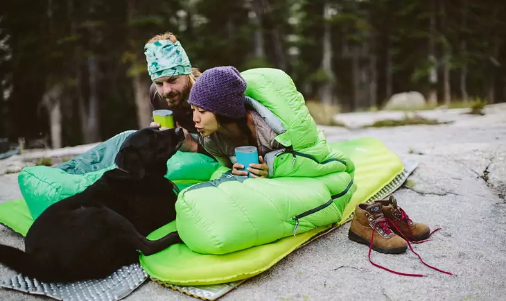 A couple in sleeping bags sitting outside with their dog